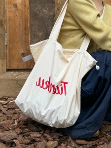 Minimalist beige nylon tote bag with red lettering and drawstring detail in outdoor setting