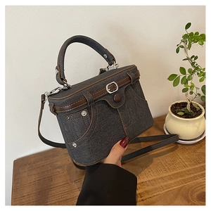 Detail of grey denim box bag with brown leather trim and silver metal rivets on wood