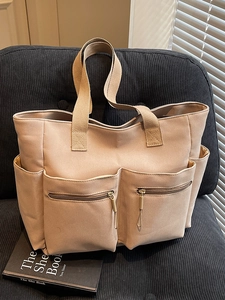 Minimalist beige canvas tote bag with multiple pockets and wide shoulder straps on a cozy chair
