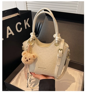 Gros plan du sac seau en cuir PU blanc crème avec bord ondulé, poignées nouées et breloque ours