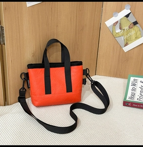 Detail einer leuchtend orangefarbenen Umhängetasche mit schwarzen Griffen und Label