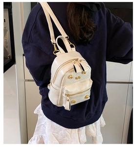 Model wearing a mini cream-white PU leather backpack with gold rivets in a casual indoor setting