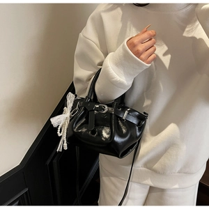Glossy black pleated bucket bag with a lace bow and pearl charm shown on a shoulder strap