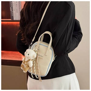 Détail du sac bandoulière beige avec motif litchi, bords bleus et breloque ours