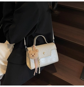 Detail view of white square bag with wavy texture, pearl buckle and brown plush bear charm