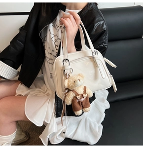 Close-up of white crossbody handbag with plush bear charm on a dark leather sofa