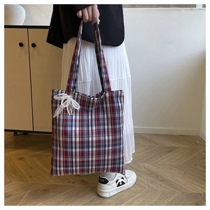 Close-up of red and blue plaid tote bag with white lace bow charm held by a person