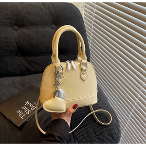 Detail view of beige shell bag with dual handles and heart pendant on a velvet sofa
