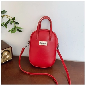 Detail of red oval crossbody bag with top handle and brand tag on dark wooden surface