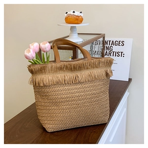 Detailed view of woven straw tote bag with fringe edge and leather handles on wood table