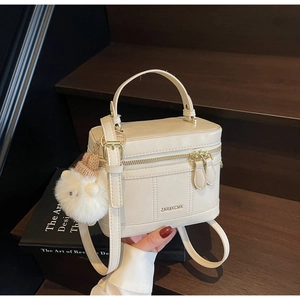 Detail view of cream leather box bag with dual zippers and plush charm on dark background