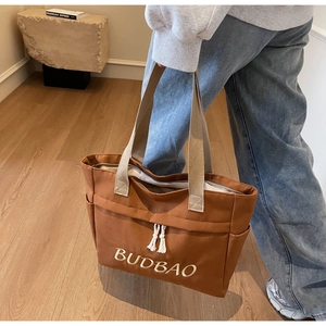 Detail of a brown canvas tote bag with embroidered letters and white tassel decoration