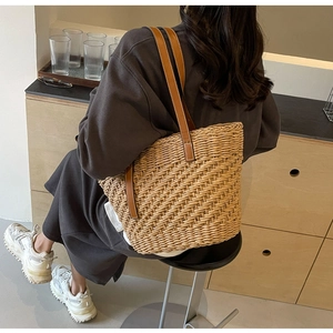 Detail of woven straw tote bag with brown leather strap worn by model in cozy interior
