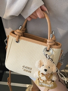 Chic beige woven bucket bag with brown leather trim and a cute teddy bear pendant
