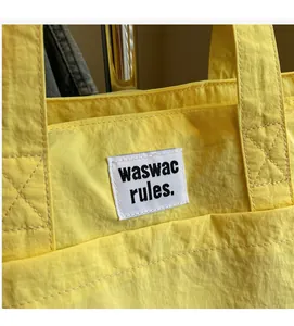 Detail of a bright yellow tote bag with textured fabric and minimalist waswac rules logo