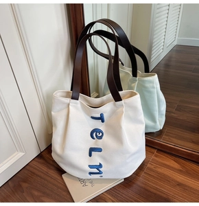 Detail of a large white canvas tote bag with brown leather straps and blue embroidery