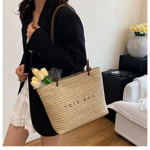 Detail of a beige woven straw tote bag with black letters carried by a model