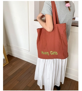 Detail of brick red canvas tote bag with green Young Girls embroidery shown on model