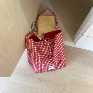 Detail of red gingham tote bag with brand label and ribbon tie closure on wood floor