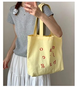 Detail of light yellow canvas tote bag with red letter print shown on shoulder
