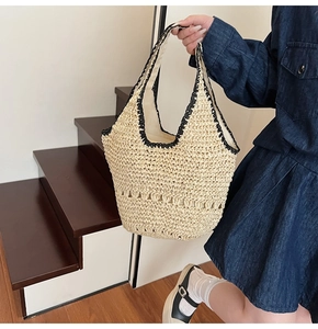 Model carrying a beige woven straw tote bag with black contrast trim on a wooden staircase