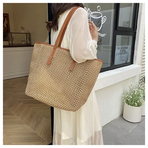 Large woven straw tote bag with leather strap handles worn over the shoulder by a model