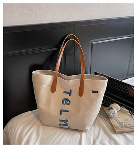 Detail of off-white canvas tote bag with brown leather straps and blue letter embroidery