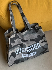 Large camouflage canvas tote bag with white GUGUGUGII 86 lettering on a cozy sofa