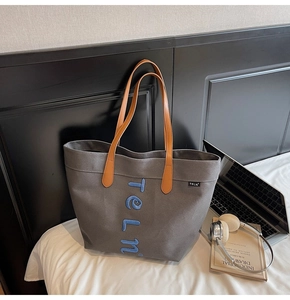Detail view of grey canvas tote bag with blue letter embroidery and brown leather handles