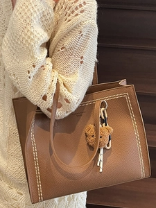 Brown leather tote bag with gold rivets and teddy charm worn by a model in casual knitwear