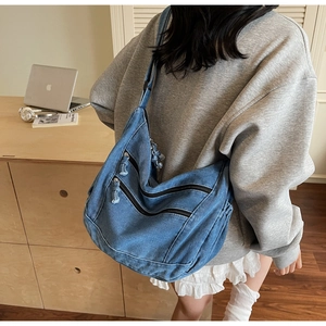 Model wearing a large denim blue crossbody bag with multiple zippers and adjustable strap