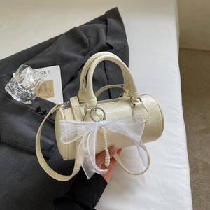 Detail of a beige cylinder handbag with oversized mesh bow and pearl chain on a white bed