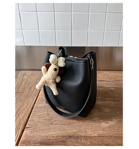 Detail of black pebbled leather bucket bag with wide strap and poodle plush charm