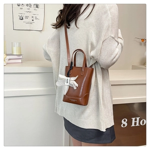 Brown leather tote bag shown with a ribbon charm on a model in a cozy interior