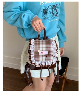 Detail eines braun-karierten Mini-Rucksacks mit weißen Spitzenrüschen und Kirsch-Patch