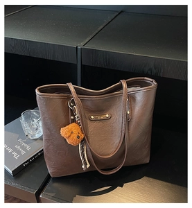 Detail of a brown tote bag featuring a metal brand plate and a cute plush bear pendant
