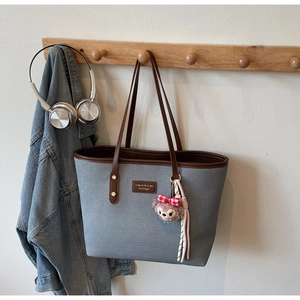 Detail of grey-blue tote bag with brown leather trim and plush doll charm hanging on a hook