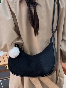 Chic black suede crescent bag with plush ball charm worn over a classic trench coat