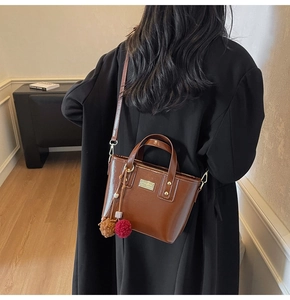 Borsa a secchiello in pelle marrone scuro con targa dorata e ciondolo in peluche su modella