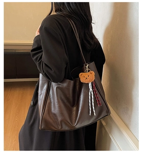 Brown leather tote bag with plush bear charm worn by a model in a cozy indoor setting