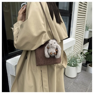 Détail du sac à bandoulière en daim marron mat avec breloque lapin en peluche sur un trench