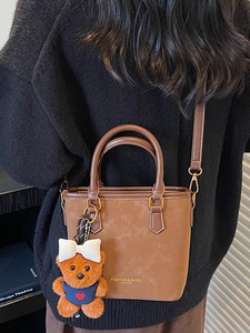 Brown leather tote bag with plush bear charm worn by a model in a casual street setting