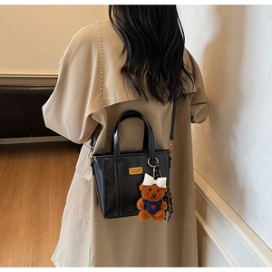 Fashionable black faux leather tote bag with plush bear charm worn by a model in soft light