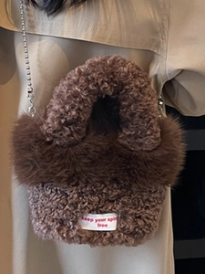 Braune Teddy-Stoff Tasche mit Metallkugelkette und weißem Etikett auf beigem Mantel
