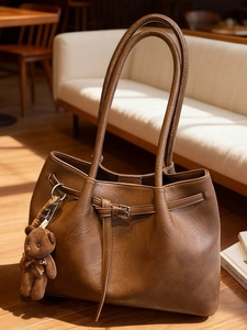 Vintage brown leather tote bag with long straps and a plush bear charm on white sofa