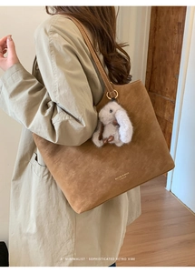Brown suede tote bag shown on shoulder featuring a gray plush bunny charm and logo