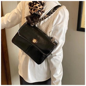 Detail of a black patent leather crossbody bag featuring gold chain and turn-lock closure