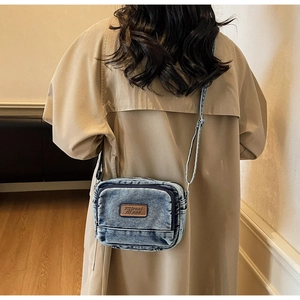 Détail d'un sac bandoulière en denim bleu avec écusson en cuir marron porté par un mannequin
