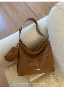 Brown leather tote bag featuring contrast stitching and a side pouch on a beige sofa