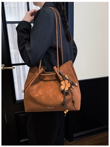 Detail of a brown suede tote bag featuring a leather floral charm and a long shoulder strap